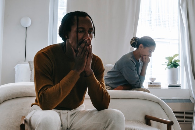 A man and a woman looking concerned, frustrated, or distraught. Their backs are to each other, sitting in a bedroom or hotel room. Perhaps they