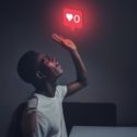 Photo of a dark skinned man holding his left palm upwards towards the celing, looking up, and a floating social media icon is sitting above it, indicating zero hearts.