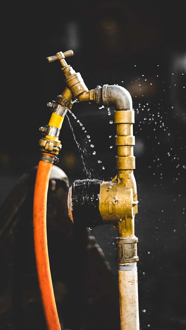 Leaking brass faucet and pipe spraying water against a dark background.